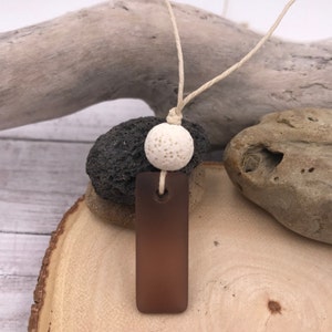 Könnte beinhalten: Ein brauner Glasanhänger mit einer weißen Lavakugel an einer natürlichen Bindfadenkordel. Der Anhänger ist rechteckig und hat eine leicht abgerundete Oberseite. Die Lavakugel ist strukturiert und hat ein Loch in der Mitte. Die Kordel ist über der Lavakugel zu einem Knoten gebunden.