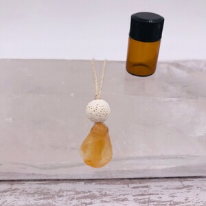 Könnte beinhalten: Eine weiße Lava-Diffusor-Halskette mit einem natürlichen orangefarbenen Citrin-Anhänger. Die Halskette ist auf einer natürlichen Bindfadenkordel aufgefädelt und wird mit einer kleinen bernsteinfarbenen Glasflasche mit ätherischem Öl geliefert.