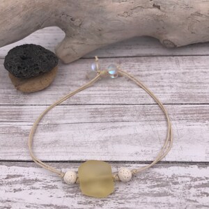 Op de afbeelding: Een armband van natuurlijke beige koord met een hanger van lichtgeel zeeglas en witte lava kralen.