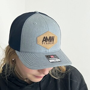 May include: A gray and black mesh trucker hat with a brown leather patch that says "AMW STABLES" in brown lettering.
