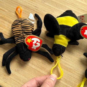 May include: Close-up of two plush toys: a spider and a bee. The spider has a striped brown and black body with black legs. The bee is black and yellow. Both toys have red heart-shaped tags.