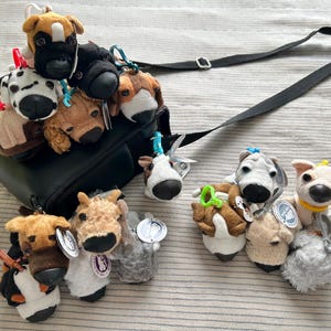 May include: A collection of plush dog keychains in various breeds and colors, featuring plastic clips for attachment. Several plushies are clustered on a black crossbody bag, while others are arranged on the surface, each with a small circular tag.