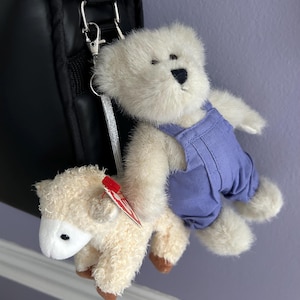 May include: A plush white teddy bear wearing blue overalls is attached to a cream-colored lamb toy. The toys are hanging from a black bag with silver hardware. The lamb has a red tag.