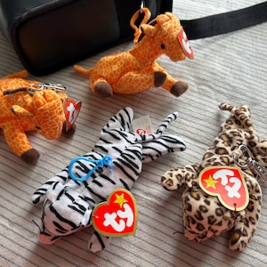 May include: Four plush animal keychains featuring two orange and yellow giraffes with brown hooves, one white and black striped zebra, and one tan and brown leopard. Each plush toy includes a red heart-shaped Ty tag and a metal or plastic clip.