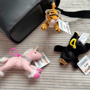 May include: Three plush animal keychains including a tiger with an orange clip, a black bear with a yellow clip, and a pink unicorn with a pink clip. Each toy features an Animal Alley tag and a Toys R Us logo, with names Sable, Cole, and Darla.