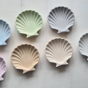 Könnte beinhalten: Sechs Keramik-Schalen in Form von Muscheln in verschiedenen Pastellfarben, darunter hellblau, grün, rosa und beige. Die Schalen sind in einer Reihe auf einer weißen Oberfläche angeordnet.