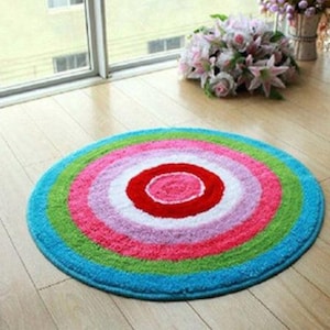May include: A round, multi-colored bath mat with a concentric circle design. The mat has a bright red, pink, white, light purple, pink, green, and blue color scheme.