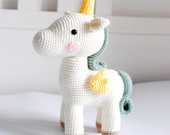 Puede incluir: Un unicornio de peluche blanco tejido a crochet con un cuerno amarillo, una melena verde y alas amarillas. El unicornio tiene pezuñas marrones y mejillas rosadas.