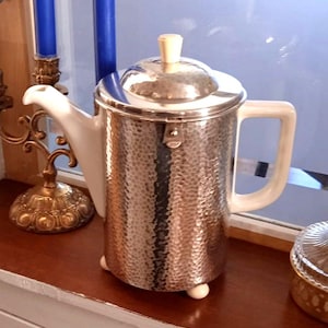 Könnte beinhalten: Eine silberfarbene, gehämmert Metall-Teekanne mit weißem Keramikausguss und Griff. Der Deckel hat einen hellfarbigen Knopf. Die Teekanne steht auf kleinen weißen Füßen. Ein dekorativer goldener Kerzenhalter und blaue Kerzen sind im Hintergrund.