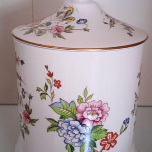 Può includere: Un barattolo in ceramica bianca con coperchio, ornato da un motivo floreale rosa, blu e rosso. Il barattolo ha finiture dorate e una forma arrotondata, per cucina o arredamento.