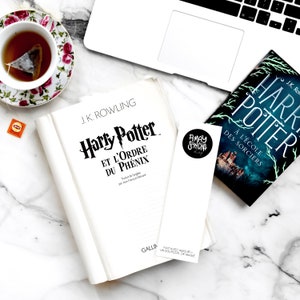 K&ouml;nnte beinhalten: Ein wei&szlig;es Buch mit dem Titel "Harry Potter und der Orden des Ph&ouml;nix" von J.K. Rowling. Das Buch liegt auf einer wei&szlig;en Marmorfl&auml;che mit einer Tasse Tee, einem Lesezeichen und einem Exemplar des Buches "Harry Potter und der Stein der Weisen" in franz&ouml;sischer Sprache.