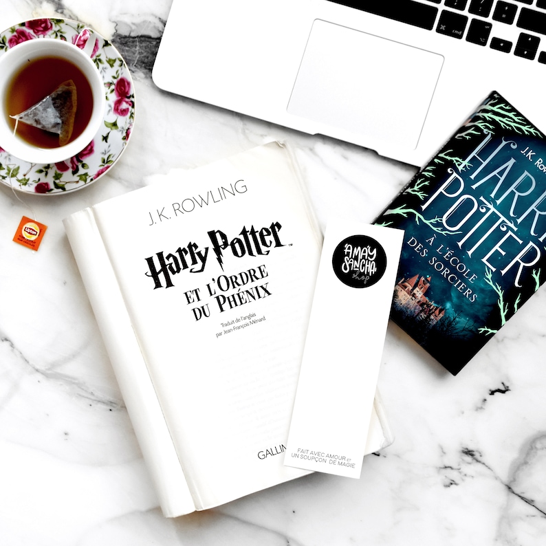 K&ouml;nnte beinhalten: Ein wei&szlig;es Buch mit dem Titel "Harry Potter und der Orden des Ph&ouml;nix" von J.K. Rowling. Das Buch liegt auf einer wei&szlig;en Marmorfl&auml;che mit einer Tasse Tee, einem Lesezeichen und einem Exemplar des Buches "Harry Potter und der Stein der Weisen" in franz&ouml;sischer Sprache.