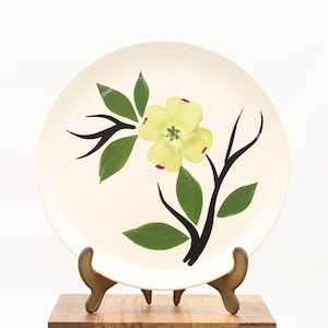 Puede incluir: Plato de cerámica color crema con diseño floral. El plato presenta una flor amarilla con hojas verdes y ramas negras. El plato se exhibe sobre un soporte de madera.