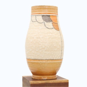 May include: A decorative ceramic vase with a cream-colored body and a textured surface. The vase features orange, gray, and black geometric patterns. The base and rim are a darker shade of orange. The vase is displayed on a wooden stand.