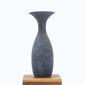 May include: A tall, blue and black speckled vase with a flared top. The ceramic vase is displayed on a small, rectangular wooden base. The vase has a slender neck and a textured surface.