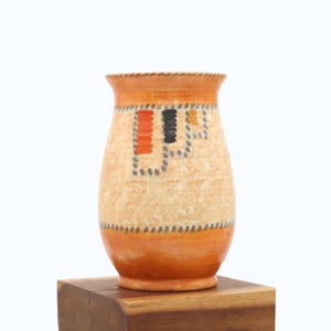 May include: A ceramic vase with a textured, speckled surface. The vase features a design of orange, black, and grey geometric shapes. The top and bottom rims are orange, and the vase is displayed on a wooden block.