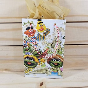 May include: A white gift bag with red and white string handles. The bag features a colorful illustration of Sesame Street characters, including Big Bird, Cookie Monster, and Elmo.