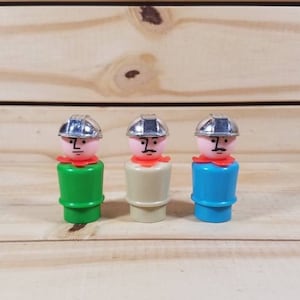 May include: Three plastic toy people wearing hard hats. The people are standing in a row on a wooden surface. The people are wearing green, beige, and blue clothing.