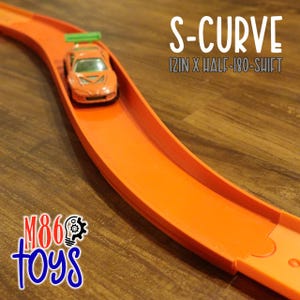 May include: An orange toy race track with an S-curve design. A small orange toy car with a green spoiler is positioned on the track. The text "S-CURVE 12IN X HALF-180-SHIFT" is displayed. The logo "M86 toys" is in the bottom left corner.