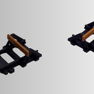 May include: Two identical black plastic track buffer stops, each featuring a rectangular orange crossbar mounted on a support frame. These components are designed with a notched base structure, showing a rigid, molded construction for model railway systems.