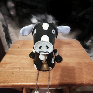 May include: A handcrafted cow figurine made from a painted terracotta pot. The cow is black with white spots and features a bell around its neck. The cow has dangling legs with decorative beads. The figurine is sitting on a wooden surface.