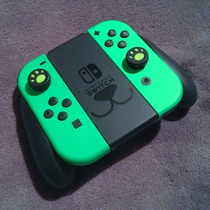以下が含まれることがあります： 緑と黒のニンテンドースイッチコントローラー。ボタンは肉球の形で、前面には肉球のデザインが施されています。コントローラーの前面には「Nintendo Switch」の文字が印刷されています。