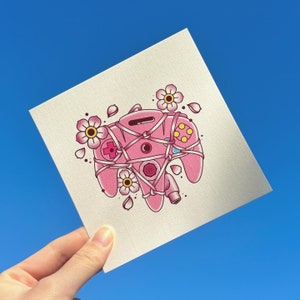 May include: A pink Nintendo 64 controller with a floral design. The controller is tied with string and has a pink and white flower design.