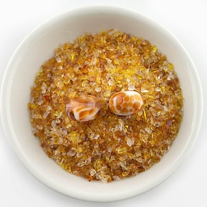 May include: A white bowl filled with amber-colored crushed glass and two decorative glass beads. One bead is white with orange swirls, and the other is white with orange spots. The glass pieces and beads are likely for crafting or jewelry making.
