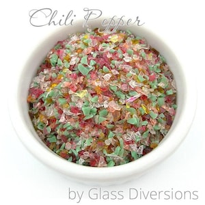 May include: A white bowl filled with colorful glass pieces in shades of red, green, and yellow. The glass pieces are small and irregular in shape, resembling crushed glass. The bowl is sitting on a white surface. The text "Chili Pepper" is written in black above the bowl, and the text "by Glass Diversions" is written below the bowl.
