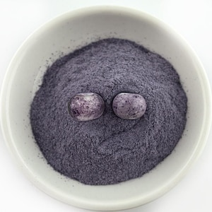 May include: A white ceramic bowl filled with a dark purple powder and two oval-shaped, speckled beads. The beads have a light purple hue with darker purple spots. The powder appears finely ground and fills the bowl.