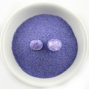 May include: A white ceramic bowl filled with fine, purple powder and two small, round, purple and white speckled beads. The beads have a smooth, polished appearance, contrasting with the textured powder. The image is brightly lit against a white background.