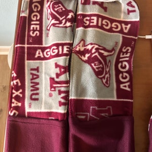 以下が含まれることがあります： グレーのフリースの裏地が付いたマルーンのフード付きスウェットシャツ。裏地には、「Aggies」と「TAMU」の文字と、マルーンと白のマスコットが繰り返しパターンで描かれています。