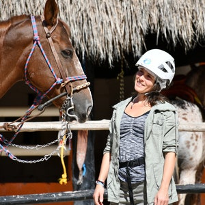 May include: A woman wearing a white helmet smiles at a brown horse with a red and blue bridle. The horse is standing in a stable with a wooden fence and a thatched roof.