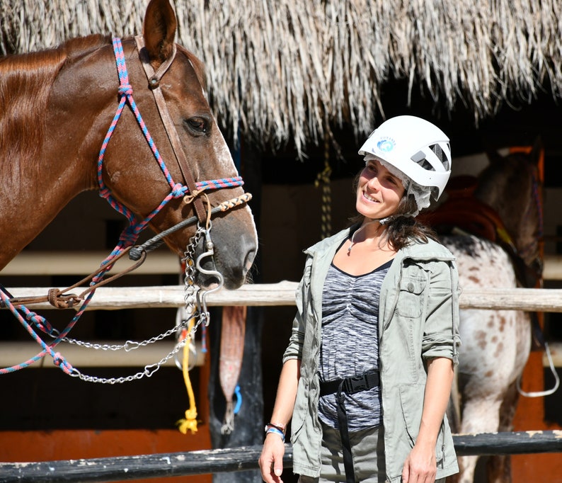 May include: A woman wearing a white helmet smiles at a brown horse with a red and blue bridle. The horse is standing in a stable with a wooden fence and a thatched roof.