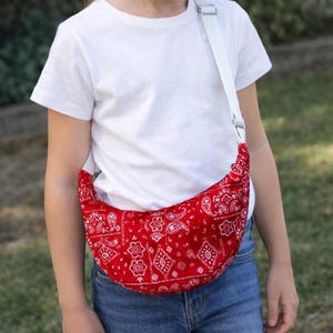 Peut inclure: Sac banane rouge à motif bandana avec une sangle blanche porté en bandoulière. Le sac en forme de croissant présente un motif cachemire blanc répété sur fond rouge. La sangle est blanche et réglable.