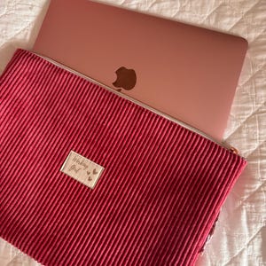 Op de afbeelding: Een donkerroze laptophoes van corduroy met een zilveren ritssluiting. Aan de voorzijde is een rechthoekige beige stoffen patch genaaid met de tekst "Working Girl" in zwarte schrijfletters, vergezeld van drie kleine hartjes op een rij.
