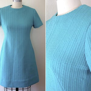 Puede incluir: Un vestido turquesa vintage con mangas cortas y cuello redondo. El vestido tiene una tela texturizada con costuras verticales y una silueta clásica en forma de A. El vestido se exhibe en un maniquí.