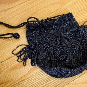 Può includere: Una piccola borsa a tracolla in perline blu scuro con chiusura a coulisse e dettaglio a frangia. La borsa ha un cordoncino nero con un fiocco.