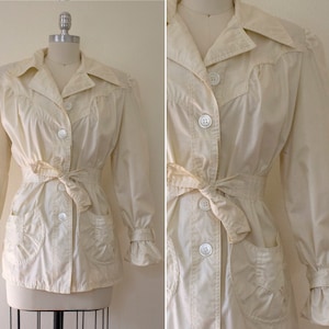 May include: A white, long-sleeved jacket with a cinched waist and a ruffled collar. The jacket has a button-up front, two pockets, and a belt.