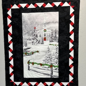 Op de afbeelding: Een gewatteerde wandhanger met een winterse kerkscène. De centrale afbeelding is een zwart-wit weergave van een met sneeuw bedekte kerk met een rode deur. De quilt is omlijst met een zwarte rand en een decoratief patroon van witte en rode driehoeken.