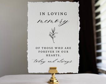 May include: A rectangular white card with deckled edges held by a gold metal stand. The card features black printed text that reads: IN LOVING memory, OF THOSE WHO ARE FOREVER IN OUR HEARTS. today and always, with a small black line drawing of a leaf.