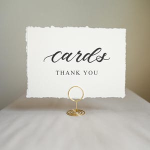 May include: A white card with the word "cards" in elegant black script, and "THANK YOU" below. The card is held by a gold wire holder, perfect for a wedding or event. The card has a deckled edge.