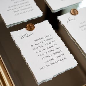 May include: Elegant wedding table setting with handmade paper place cards. Each card features a torn edge, calligraphy names, and a wax seal. The cards are arranged on a reflective surface within a gold frame.