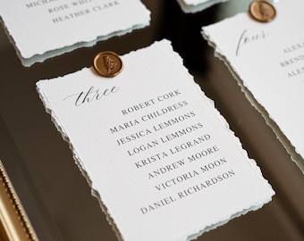 May include: A gold-framed mirror displaying multiple white paper cards with deckled edges. Each card features a table number in script, a list of names in black text, and a small circular bronze wax seal with a botanical imprint at the top center.