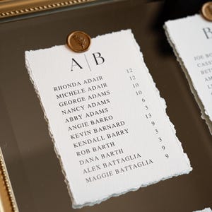 May include: Elegant wedding seating chart with names and table numbers printed on textured paper. Each card is adorned with a gold wax seal. The names are listed in a simple, classic font.