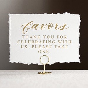 May include: A white card with gold lettering that reads "favors" in cursive. The card also says "Thank you for celebrating with us. Please take one."