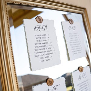 May include: A gold framed mirror displays seating assignment cards for an event. Each card is white with a deckled edge, held in place by a bronze-colored seal. The cards feature names and table numbers.