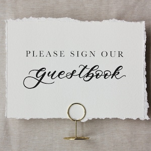 May include: A white card with the text "PLEASE SIGN OUR Guestbook" in black script. The card has a torn edge and is held by a gold-colored metal holder. The background is a light beige fabric.