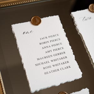 May include: Elegant wedding table setting with handwritten guest names on textured white paper. Each card is sealed with a gold wax stamp featuring a leaf design. The cards are displayed in a gold frame.