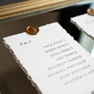 May include: A white paper place card with torn edges, featuring the word "one" in elegant script and a list of names. A gold wax seal with a floral design adorns the top. The card is set on a reflective surface.